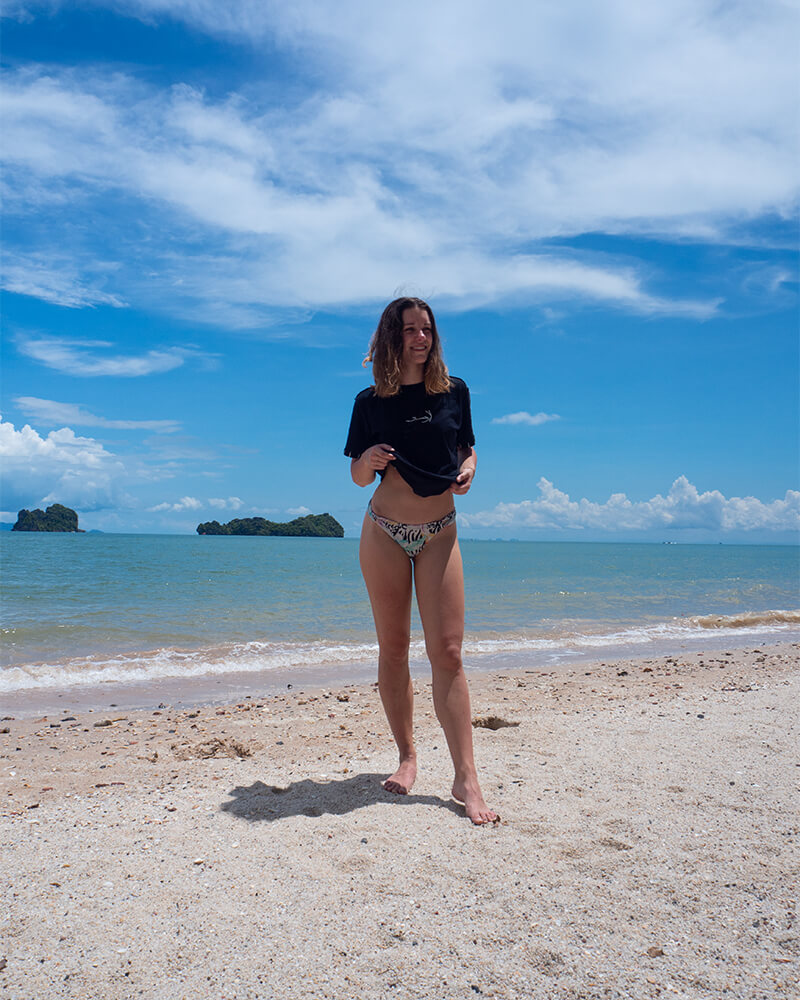 Frau mit T-Shirt und Bikini steht am Strand mit blauem Meer i
