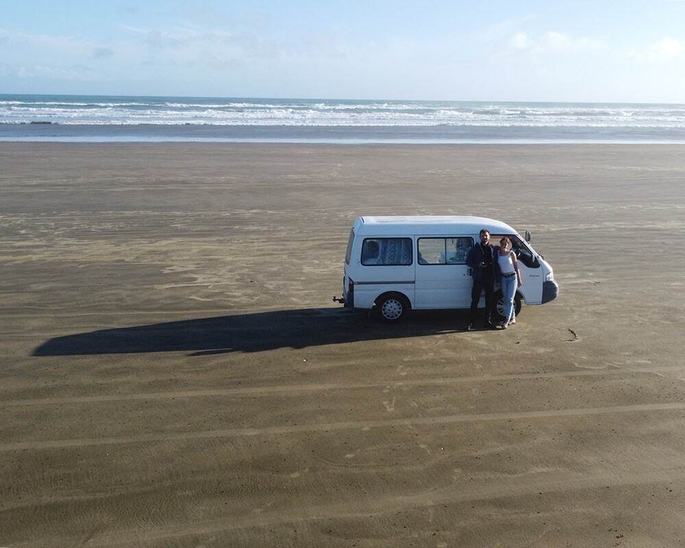 ein Junges paar steht vor einem Campervan auf einem Strand 