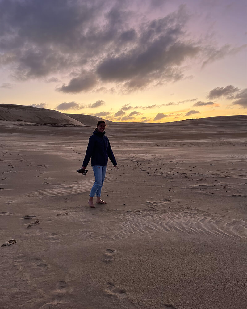 Junge Frau steht barfuß auf Sanddünen, während die Sonne untergeht