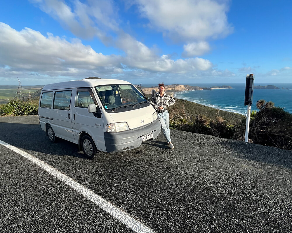 eine junge Frau steht auf einer Straße und lehnt an einem Campervan, im Hintergrund sieht man das Meer und die K+ste