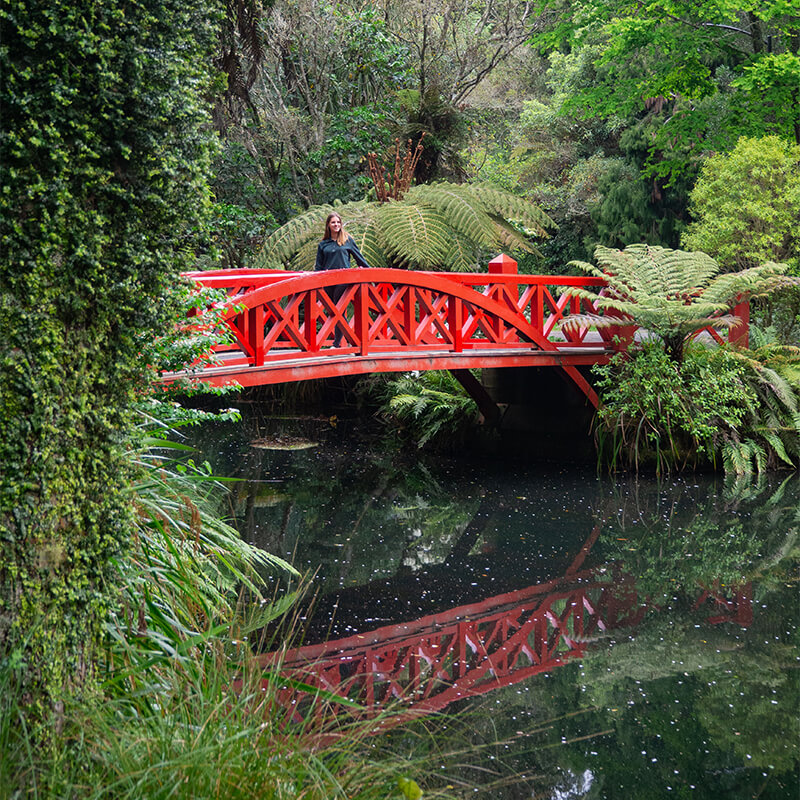 Junge Frau auf einer roten Brücke, die über einen Fluss führt der von Bäumen umgeben ist