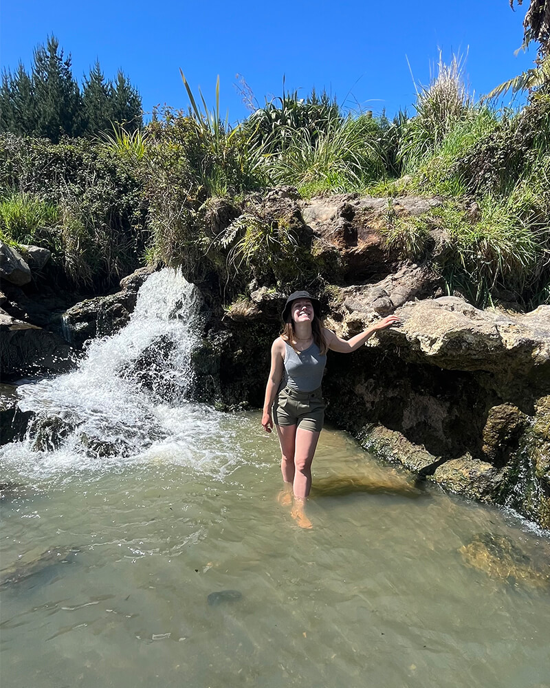 junge Frau steht in einem kleinen Wasserbecken, in das ein Wasserfall mündet