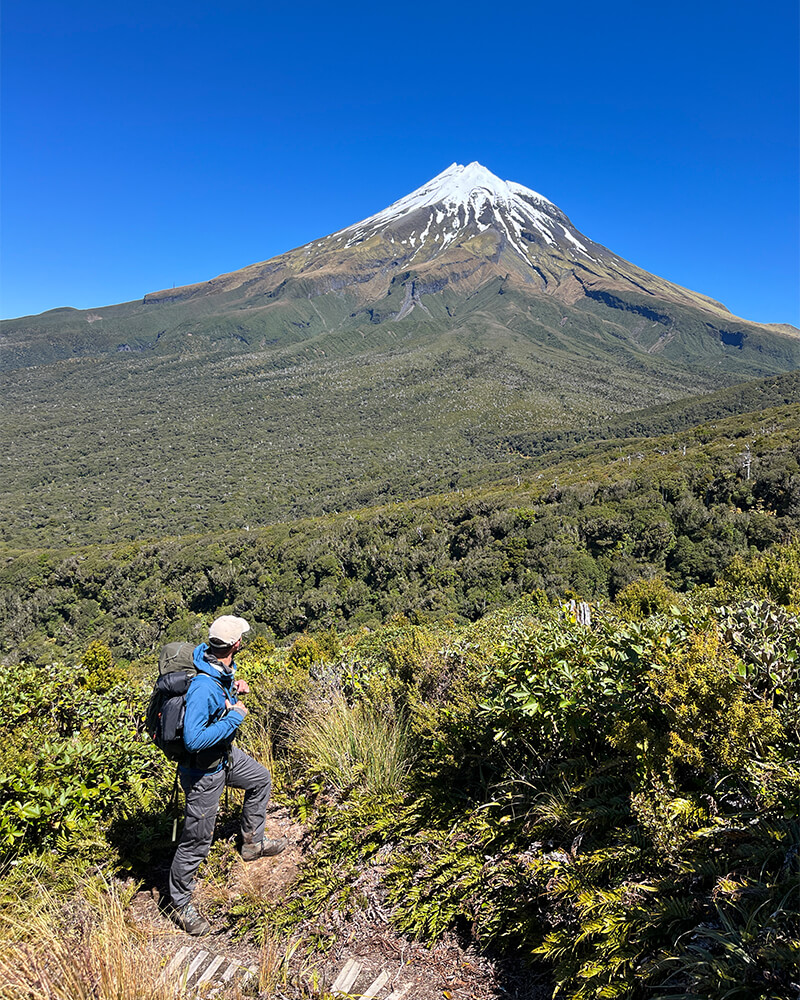 ein junter Mann mit Rucksack steht auf einem Weg mit dem Mount Taranaki im Hintergrund