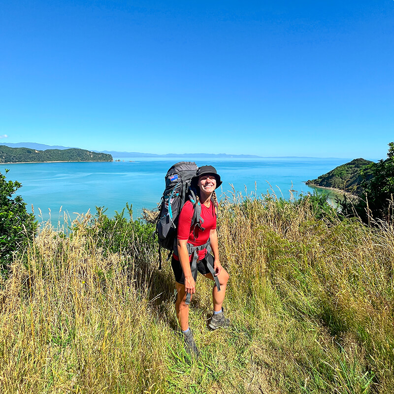 junge Frau mit großem Rucksack steht auf einer Wiese, im Hintergrund ist das Meer