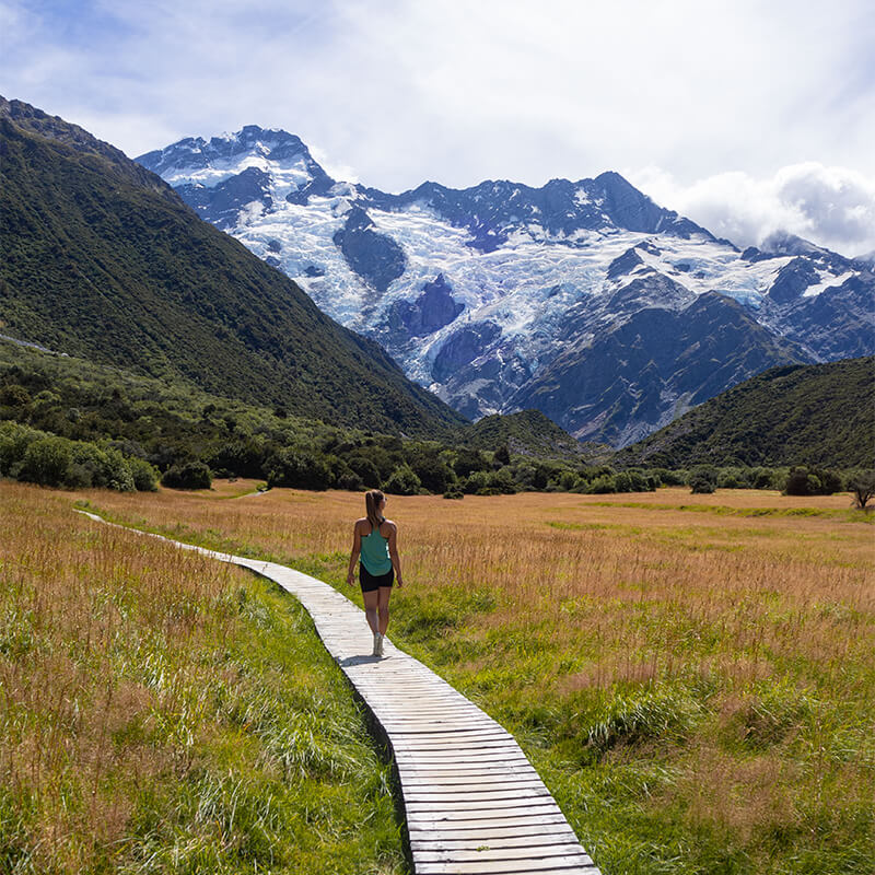 junge Frau auf Weg durch eine Graslandschaft mit einem Gletscher im Hintergrund