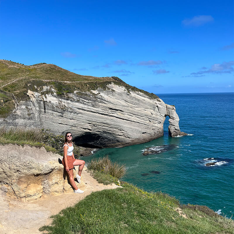 junge Frau lehnt an einem Felsen an einer Klippe, im Hintergrund ist das Meer und ein größerer Felsen