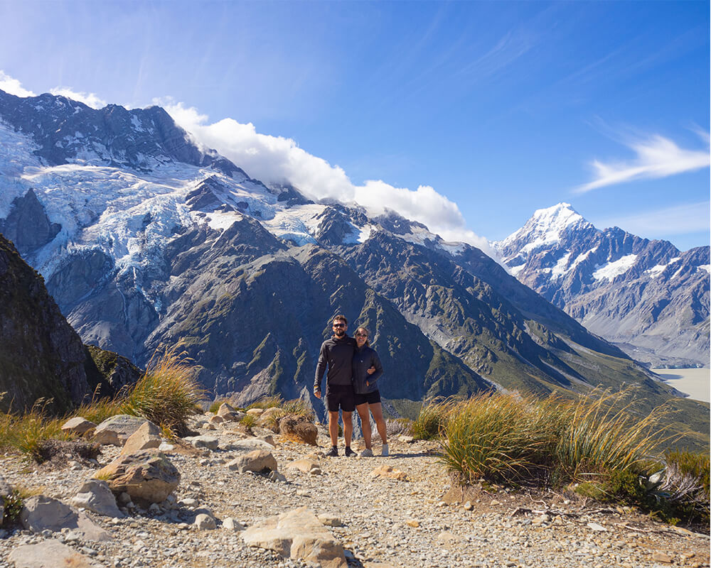 junges Paar steht auf einem Berg mit Gletscher und Mout Cook im Hintergrund