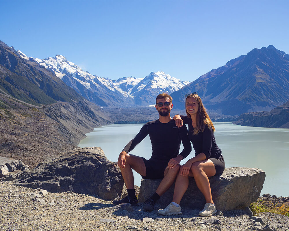 junges Paar sitzt auf einem Stein vor einem Gletschersee