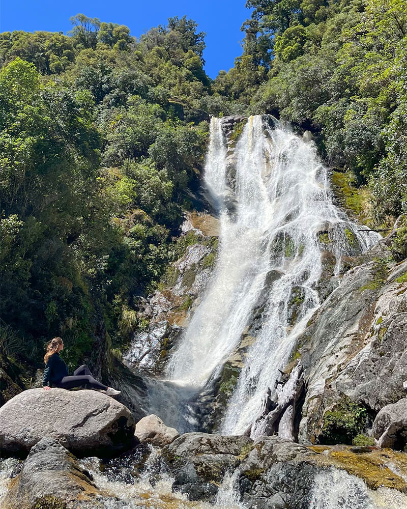 junge Frau sitzt auf einem Felsen vor einem Wasserfall