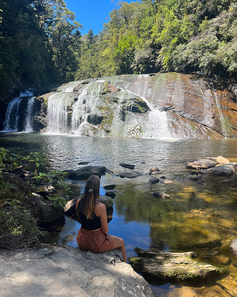 junge Frau sitzt auf einem Felsen an einem Fluss, im Hintergrund ist ein kleiner Wasserfall