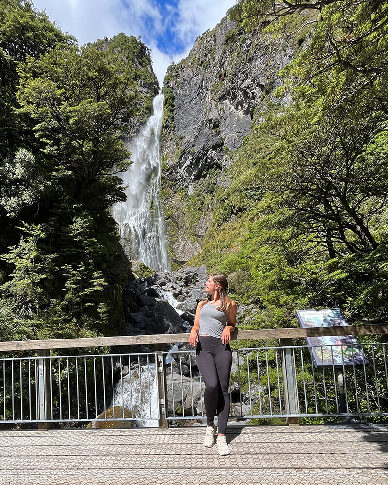 Junge Frau lehnt an einem Geländer, im Hintergrund ist ein Wasserfall
